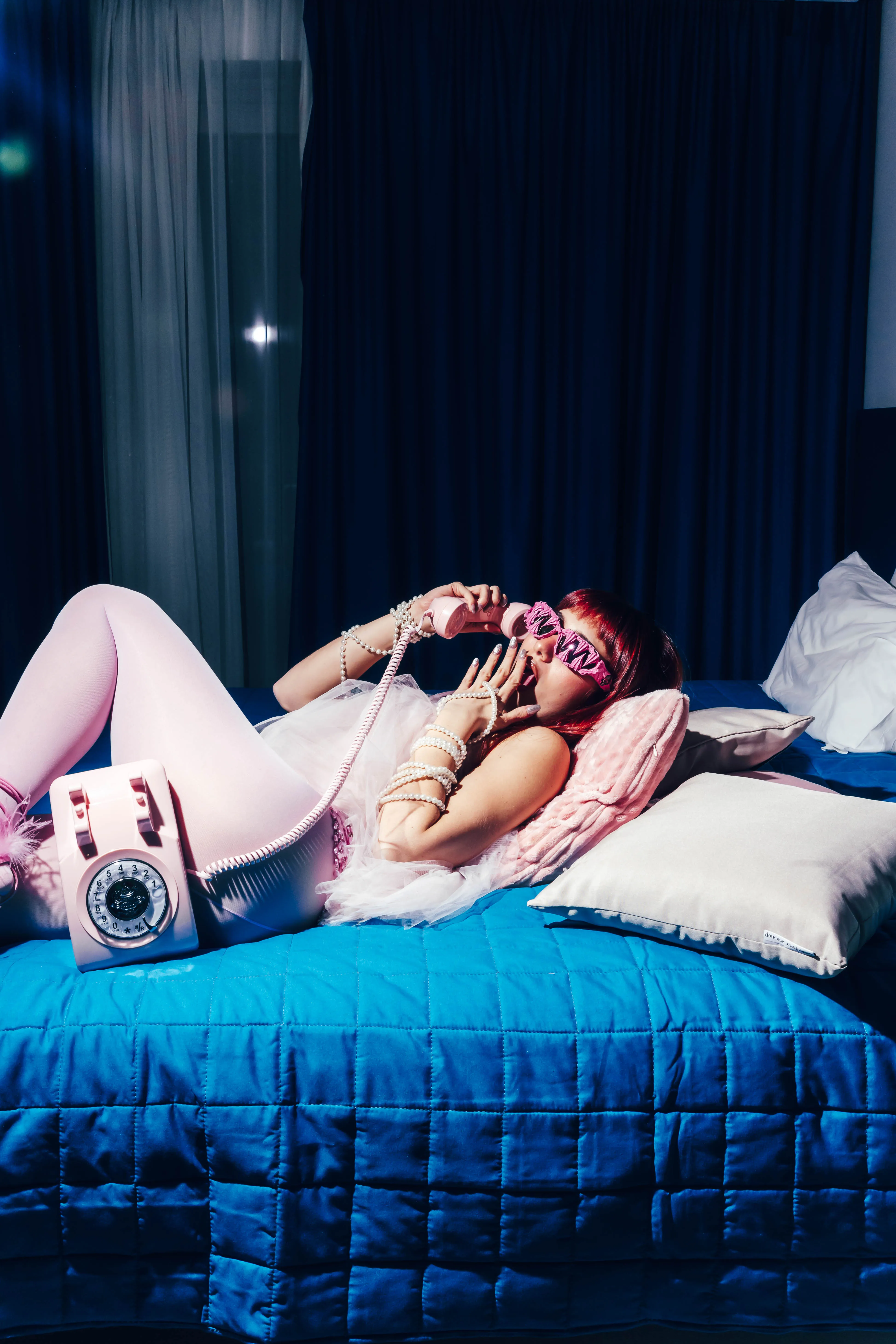 Model lounging on a blue bed with a pink rotary telephone